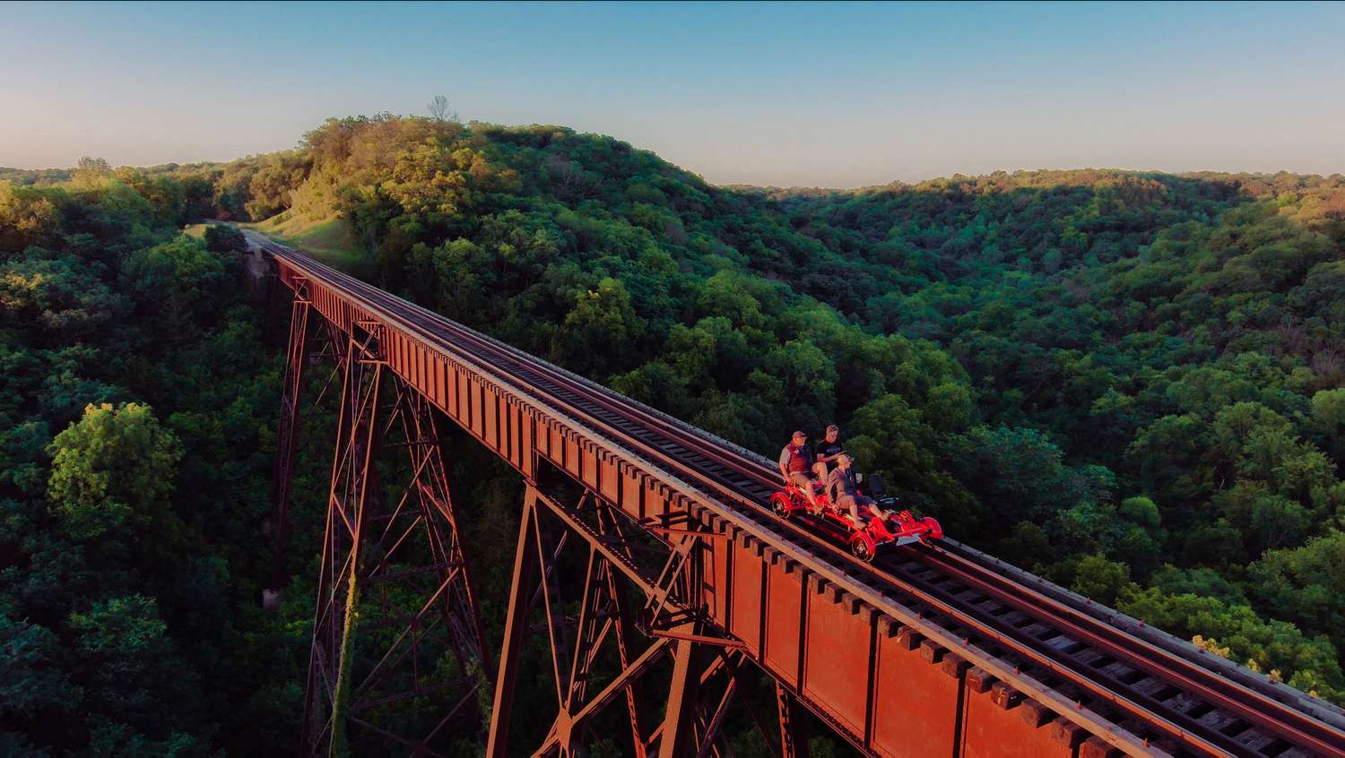 A pedal-powered car crosses a high bridge over a forested valley.